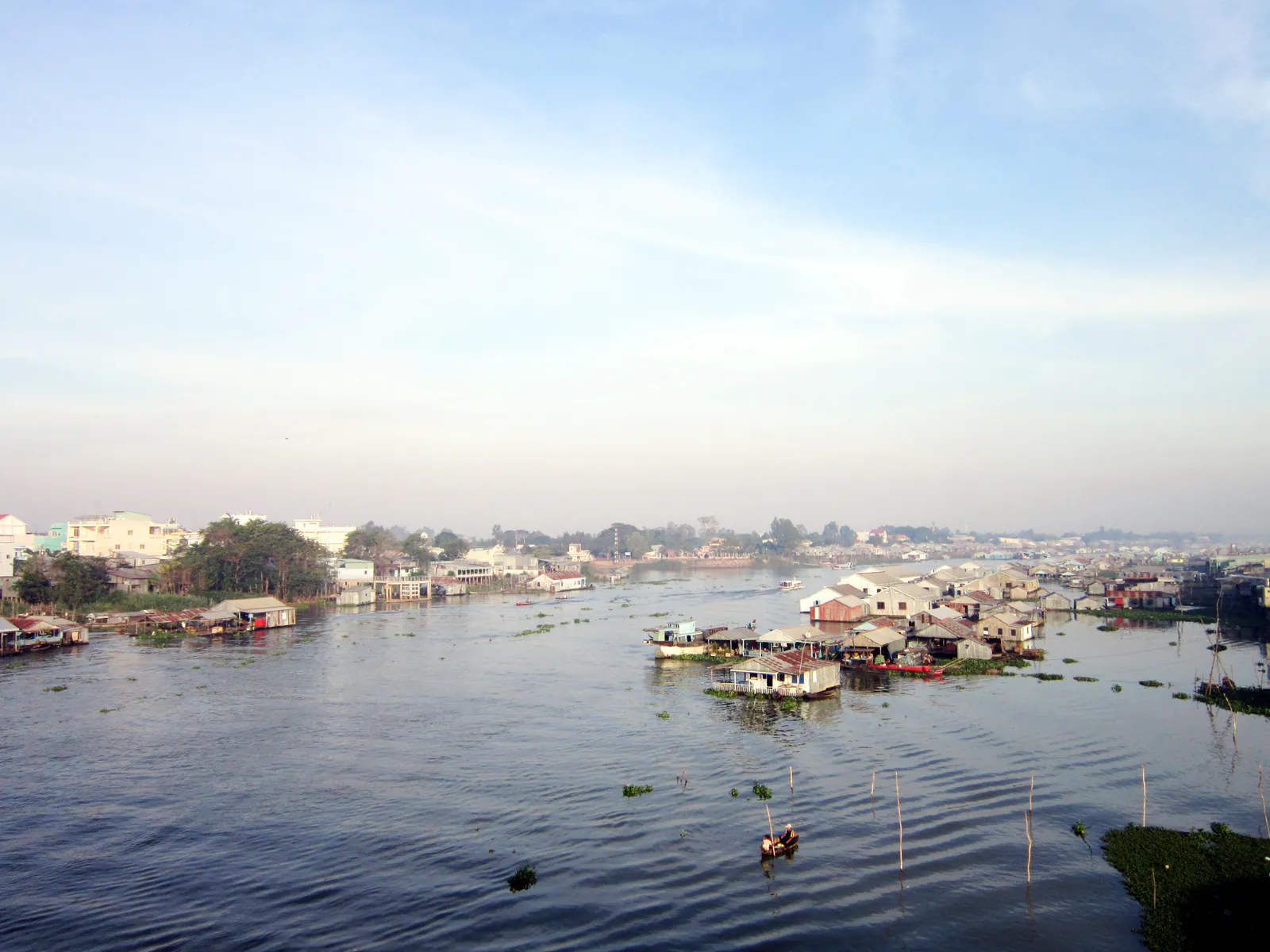 Khúc sông rộng vùng đồng bằng sông Cửu Long lúc sáng sớm, một bên là dãy nhà bè nuôi cá mái tôn nổi san sát trên mặt nước, bên kia là thị trấn thấp tầng nép sau hàng cây, một chiếc xuồng nhỏ đi giữa dòng.