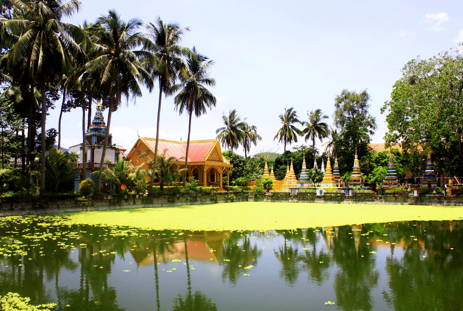 Khuôn viên chùa Xà Tón (Xvayton), Tri Tôn, An Giang nhìn qua hồ nước phủ bèo: nhà giảng mái đỏ với hàng cột chạm khắc thếp vàng, bên phải là dãy tháp mộ Khmer nhiều màu, xung quanh là rặng dừa.